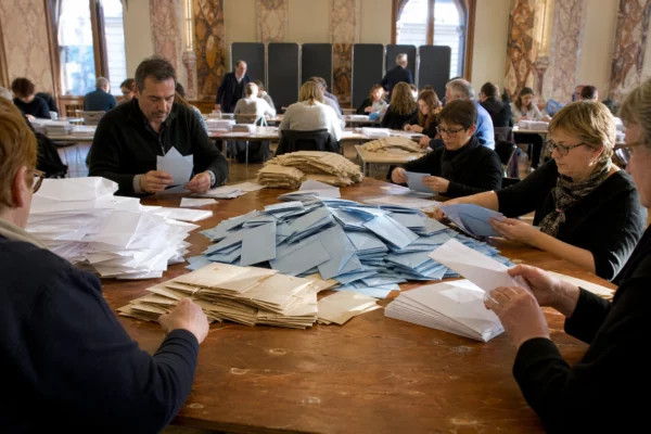 Bulletins et enveloppes de vote sur une table, dans une salle municipale où des grands électeurs préparent le scrutin.