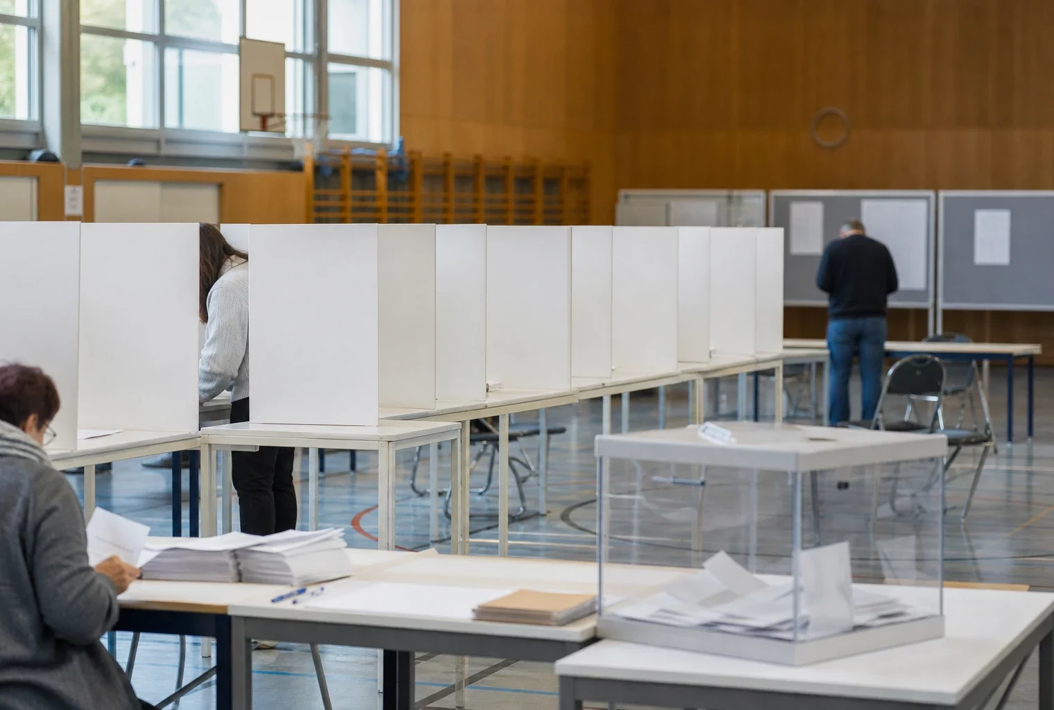 Bureau de vote allemand dans une salle lumineuse, isoloirs alignés, urne transparente et table de vote au premier plan.
