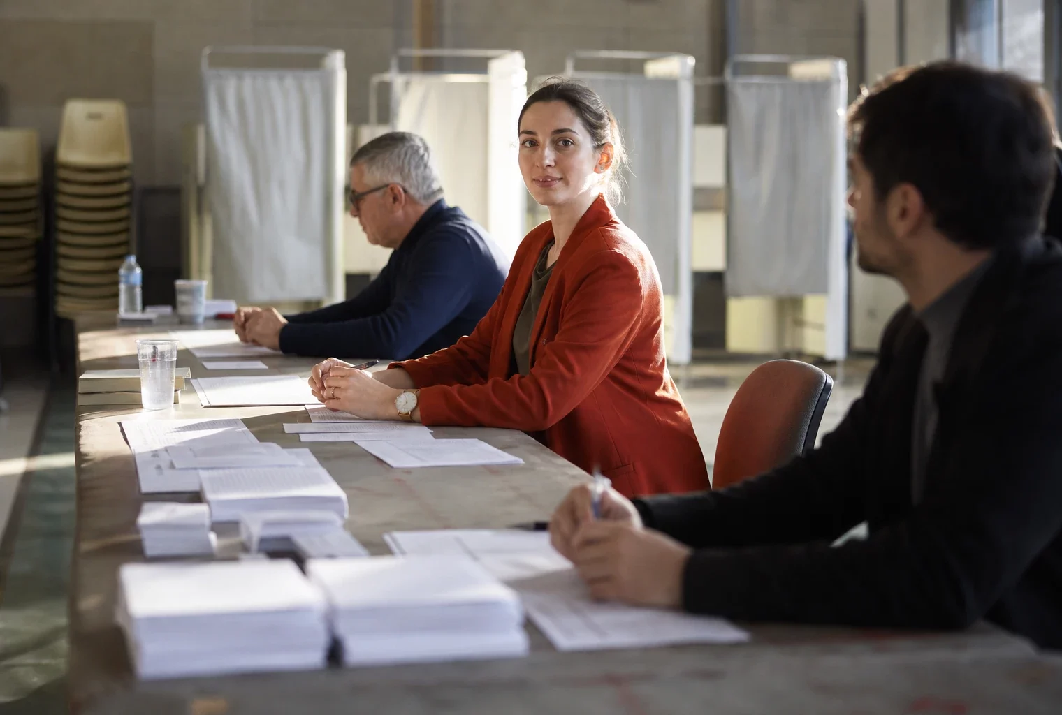 Bureau de vote de la primaire socialiste, avec bulletins, tables d’émargement et assesseurs sous une lumière dure.