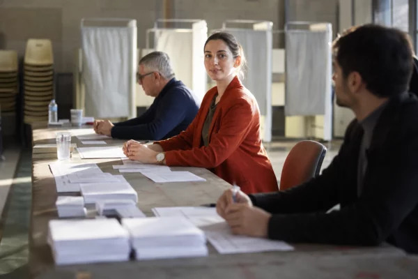 Bureau de vote de la primaire socialiste, avec bulletins, tables d’émargement et assesseurs sous une lumière dure.