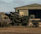 Canon d’artillerie sur base militaire, caisses de munitions au premier plan, hangar et véhicules en arrière-plan.