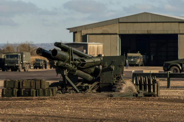 Canon d’artillerie sur base militaire, caisses de munitions au premier plan, hangar et véhicules en arrière-plan.