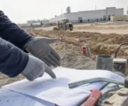 Mains gantées au premier plan sur un chantier industriel, avec engins, tranchées et hangars en arrière-plan.