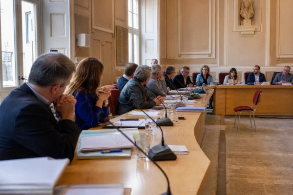 Salle de conseil municipal française avec élus anonymes, dossiers budgétaires, micros et une chaise vide