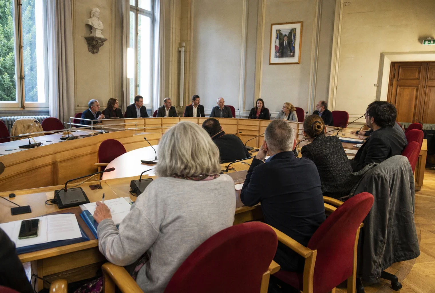 Salle municipale française avec élus anonymes, micros et chaise vide lors d’une réunion politique locale