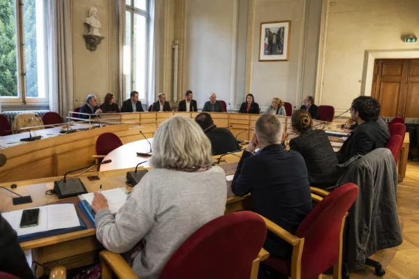 Salle municipale française avec élus anonymes, micros et chaise vide lors d’une réunion politique locale