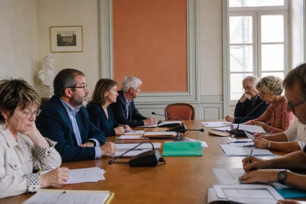 Salle de conseil municipal française avec une chaise vide, des micros et des dossiers sous une lumière naturelle claire