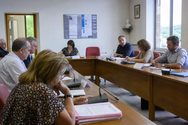 Salle de conseil municipal française avec une chaise vide, des micros et des dossiers autour d’une table