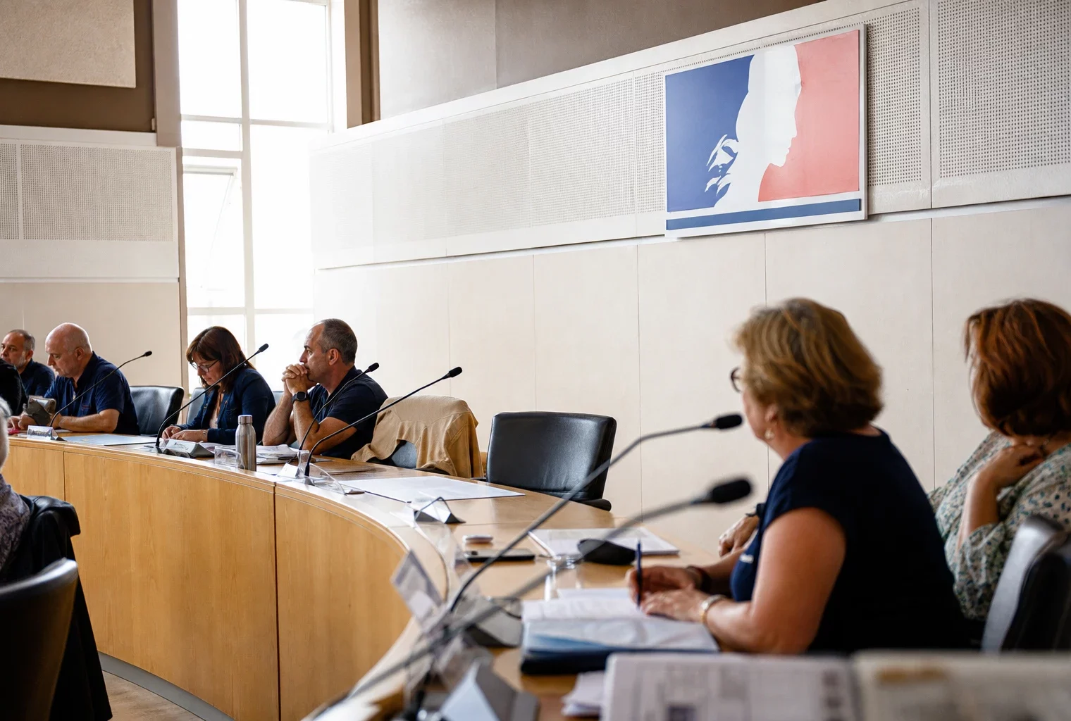 Salle de conseil municipal française avec une chaise vide, des micros et des dossiers lors d’une réunion locale