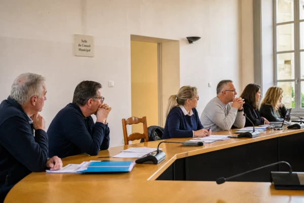 Salle municipale française avec élus anonymes, chaise vide, micros et dossiers lors d’un conseil local en Gironde