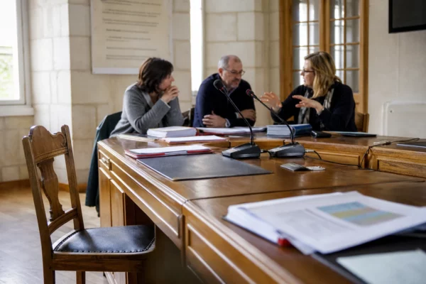 Salle municipale française avec micros, dossiers flous et chaise vide lors d’une réunion sur les menaces hybrides