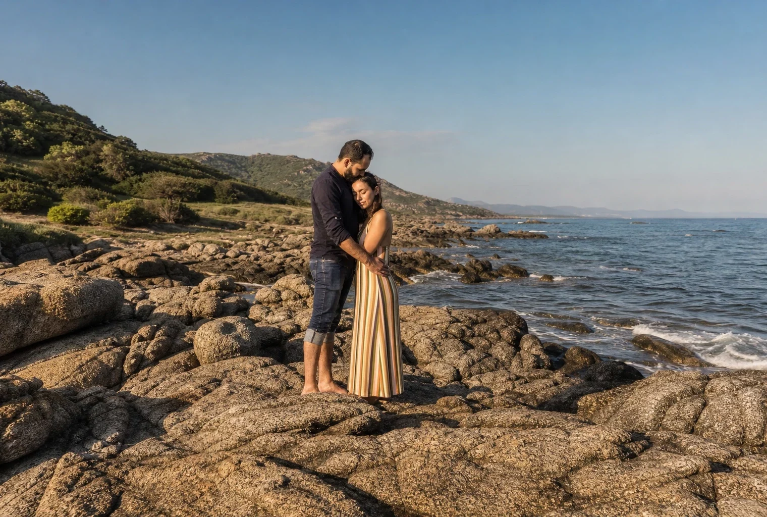 Couple enlacé sur des rochers en Corse, mer calme derrière eux et végétation méditerranéenne floue au loin.