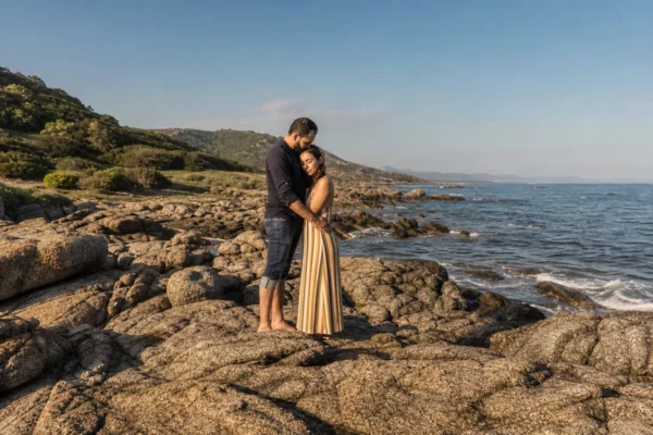 Couple enlacé sur des rochers en Corse, mer calme derrière eux et végétation méditerranéenne floue au loin.