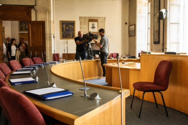 Salle municipale française préparée pour un débat politique, avec micros, dossiers et une chaise vide
