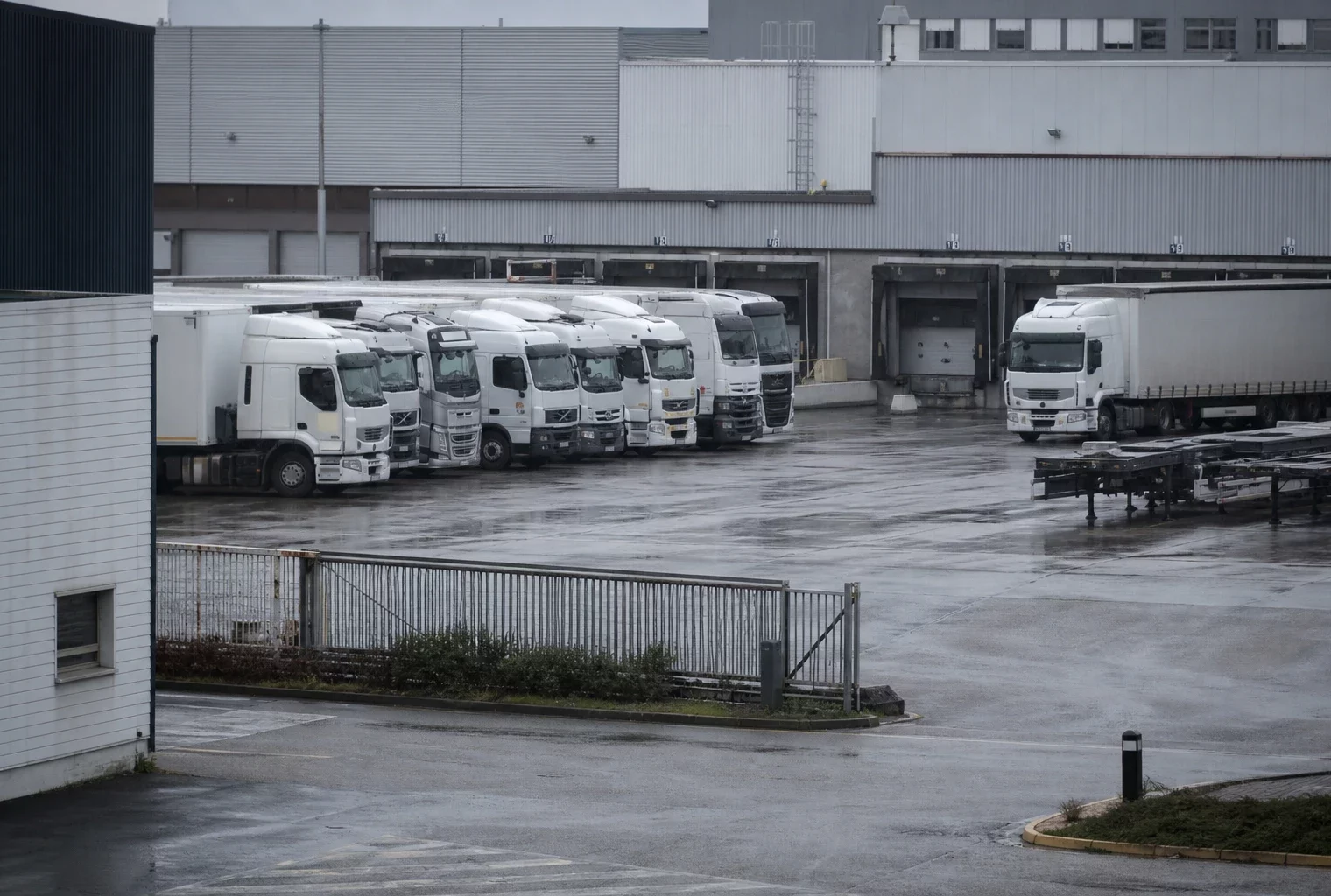 Dans un dépôt de transport routier, plusieurs camions stationnés devant un hangar logistique et des quais de chargement.