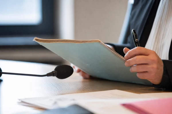 Mains anonymes tenant un dossier lors d’une audition, avec micro de commission et documents flous sur une table en bois