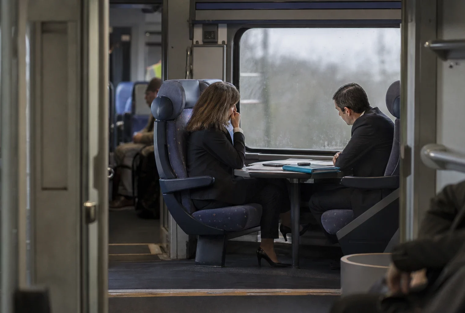 Dans un train, deux personnes échangent autour de documents posés sur une tablette.
