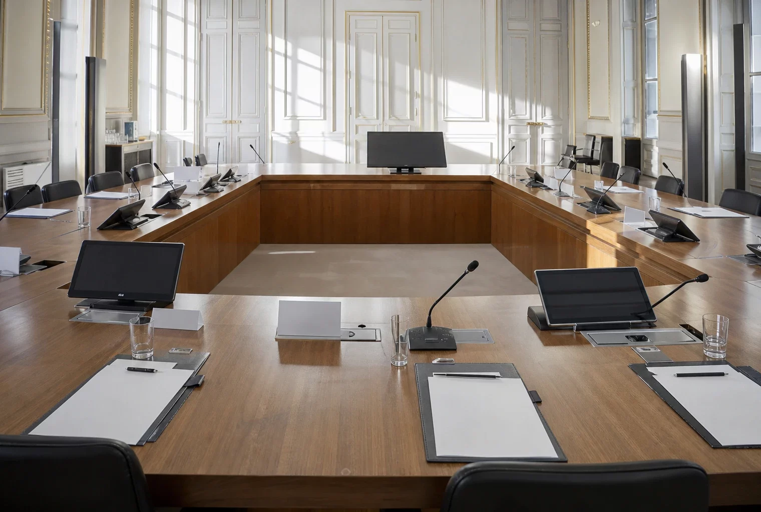 Salle de conférence de l’Élysée vide, table en U, micros et dossiers vierges sous une lumière matinale claire.