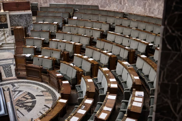 L’hémicycle de l’Assemblée nationale vu en plongée, rangées de sièges usés et pupitres sous une lumière froide uniforme.