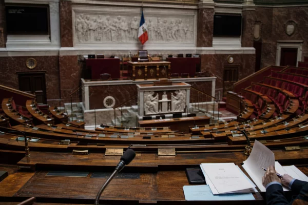 Hémicycle de l’Assemblée nationale vide, pupitres et sièges usés sous une lumière grise et pluvieuse, avec des mains au bord du cadre.
