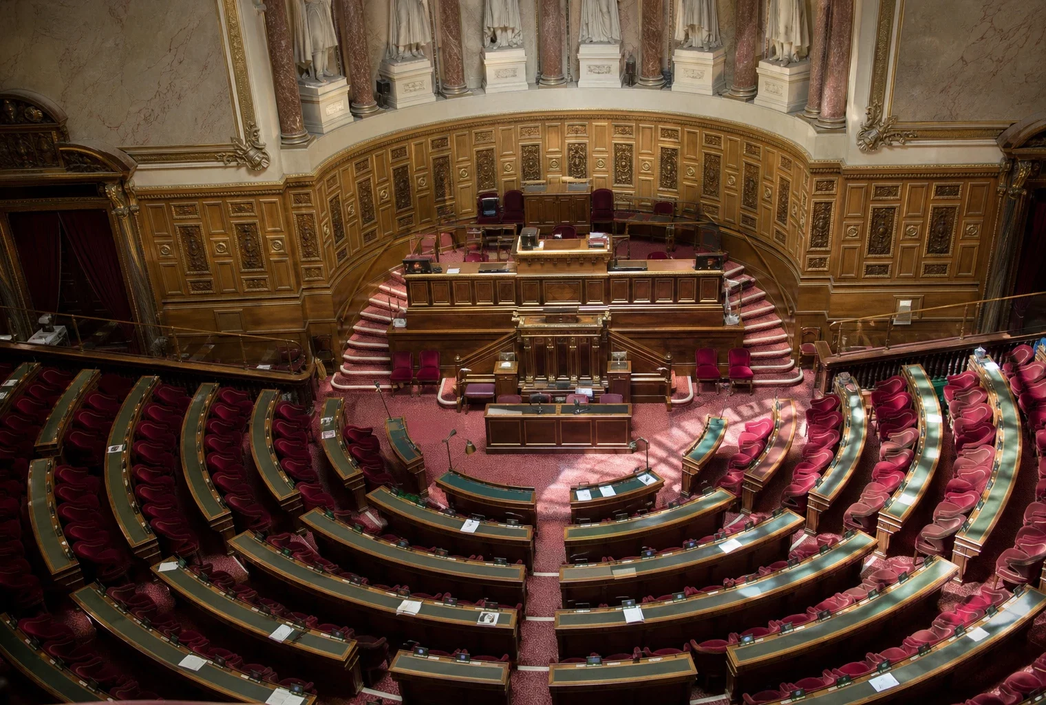 Hémicycle du Sénat vide à Paris, rangées de sièges rouges et pupitres vus en plongée, lumière de matinée nette.