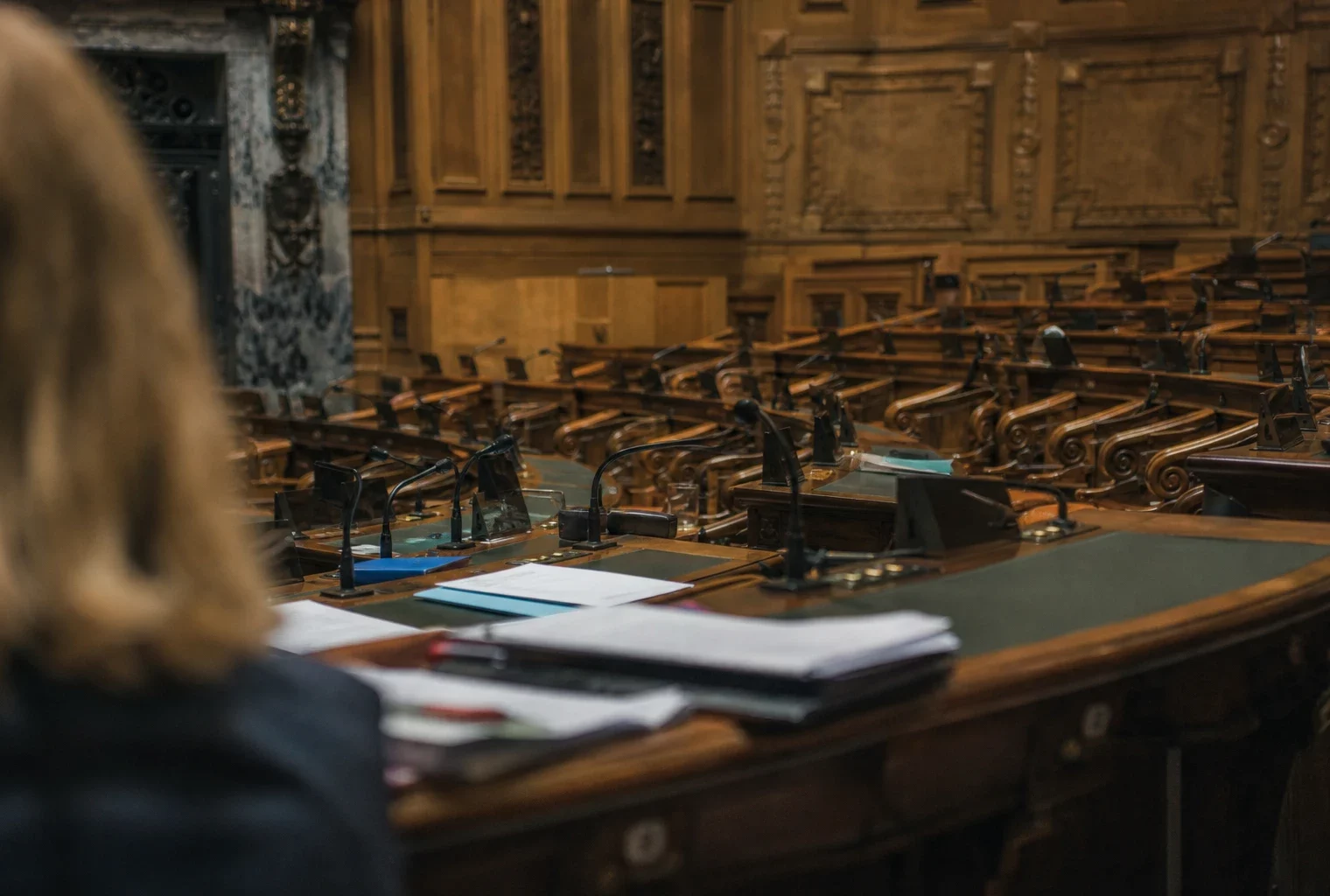 L’hémicycle du Sénat en vue oblique, pupitres et micros au premier plan, sièges de bois en profondeur sous une lumière grise.