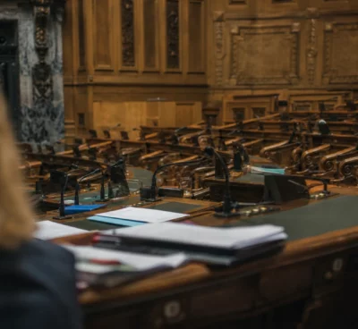 L’hémicycle du Sénat en vue oblique, pupitres et micros au premier plan, sièges de bois en profondeur sous une lumière grise.