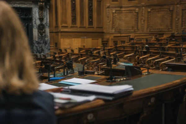 L’hémicycle du Sénat en vue oblique, pupitres et micros au premier plan, sièges de bois en profondeur sous une lumière grise.