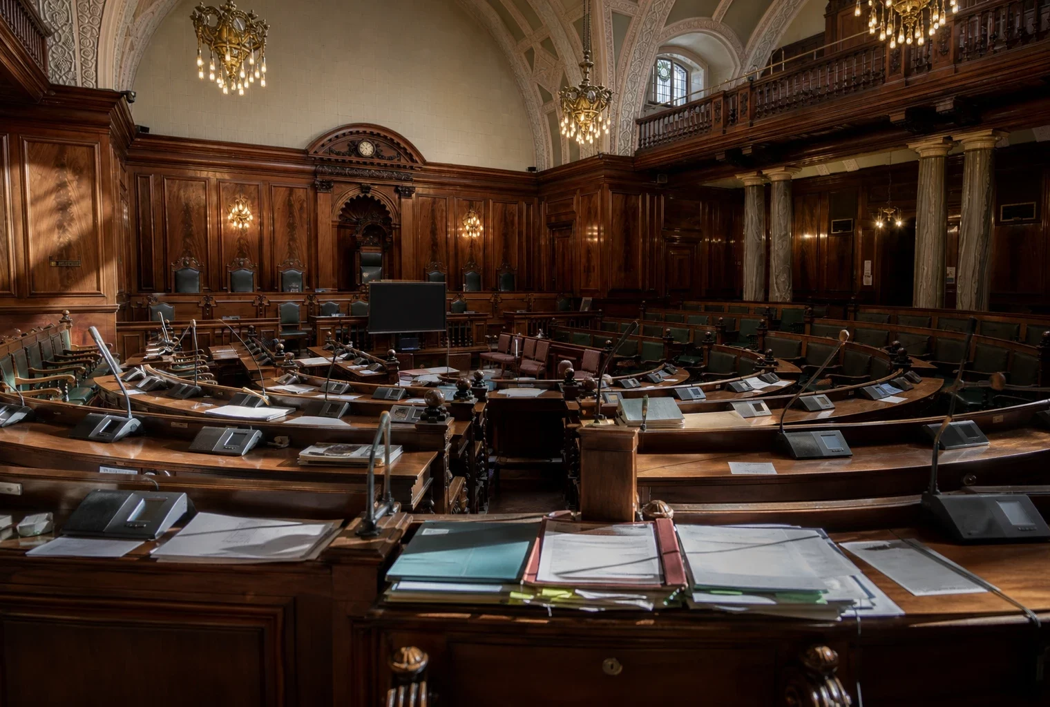 Hémicycle du Sénat vide, pupitres et micros au premier plan, boiseries et travées courbes baignées de lumière dorée.