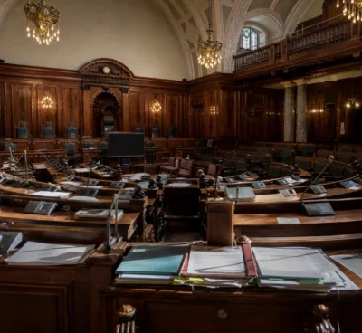 Hémicycle du Sénat vide, pupitres et micros au premier plan, boiseries et travées courbes baignées de lumière dorée.