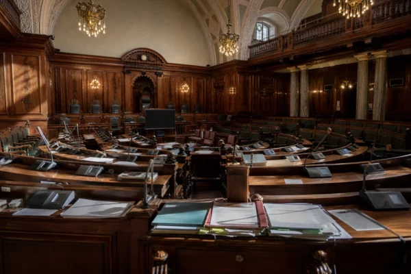 Hémicycle du Sénat vide, pupitres et micros au premier plan, boiseries et travées courbes baignées de lumière dorée.