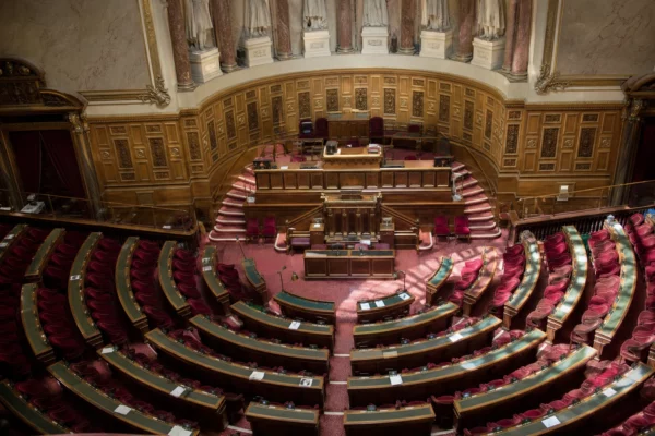 Hémicycle du Sénat vide à Paris, rangées de sièges rouges et pupitres vus en plongée, lumière de matinée nette.