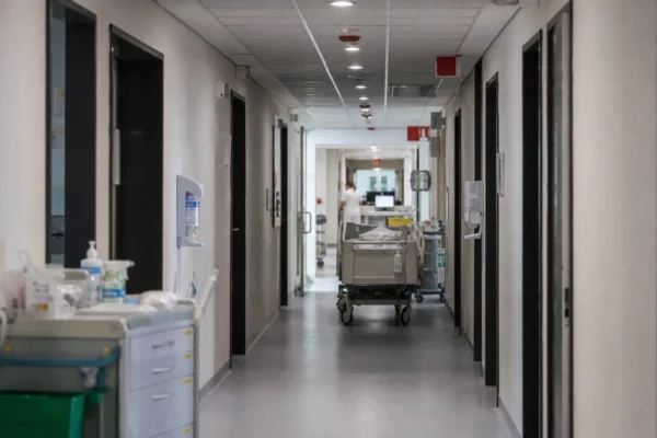 Couloir d’hôpital public avec chariot de soins, portes ouvertes sur des chambres et postes infirmiers en profondeur.