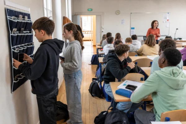Des lycéens déposent leurs téléphones dans un casier mural, sous le regard d’un enseignant, dans une classe française lumineuse