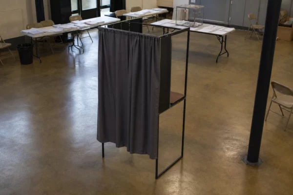 Un isoloir vide dans un bureau de vote, avec l’urne et les tables d’émargement en arrière-plan.