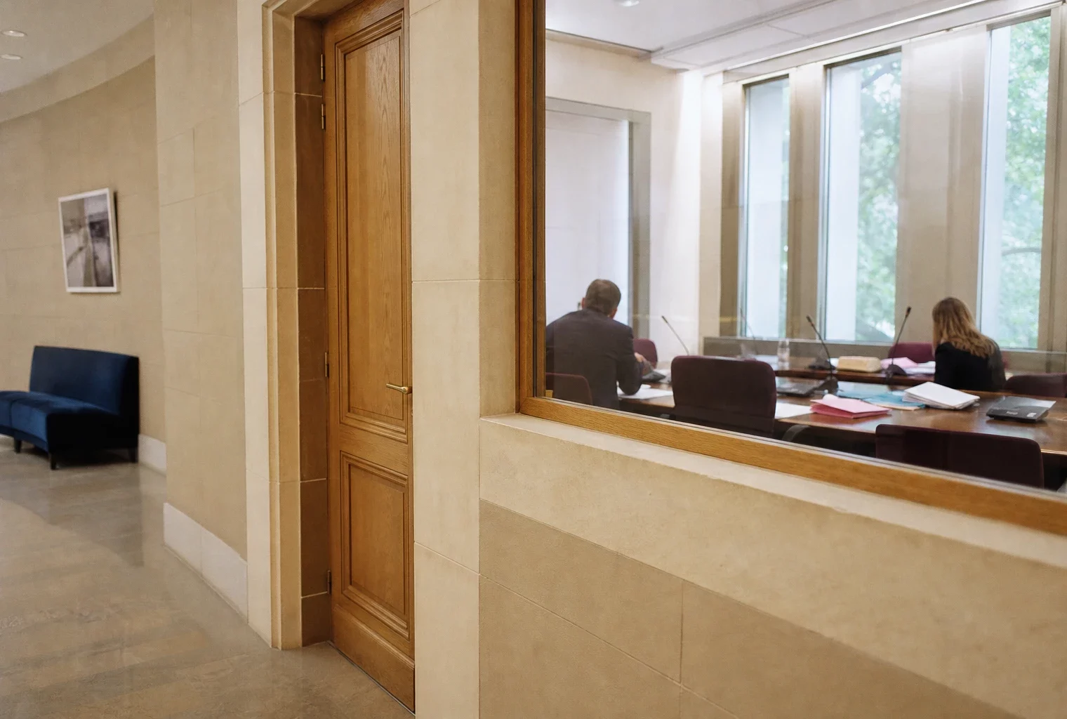 Couloir clair d’une institution publique française ouvrant sur une salle d’audition avec dossiers et micros flous