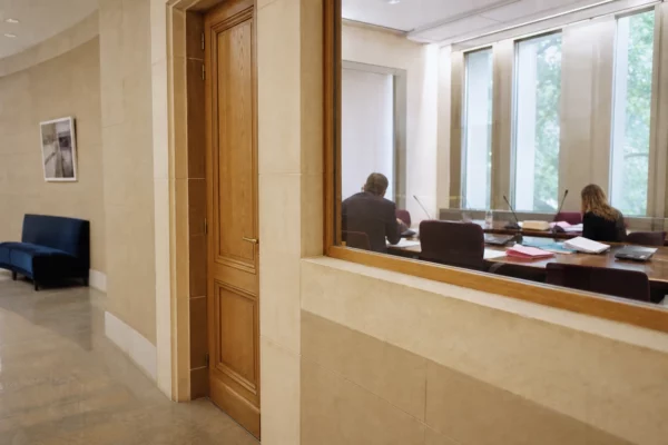 Couloir clair d’une institution publique française ouvrant sur une salle d’audition avec dossiers et micros flous