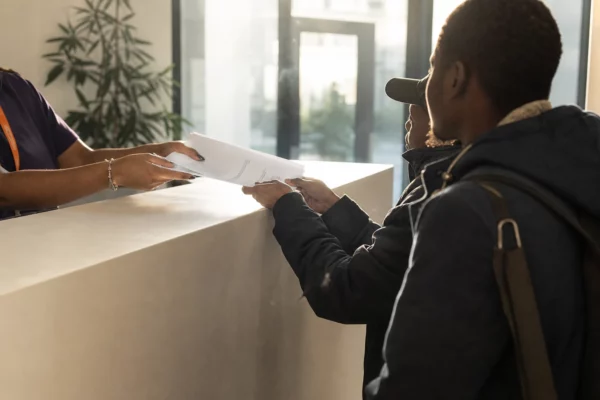 Comptoir d’accueil à la Maison Bakhita, mains échangeant des documents, personnes en attente et porte vitrée en arrière-plan.