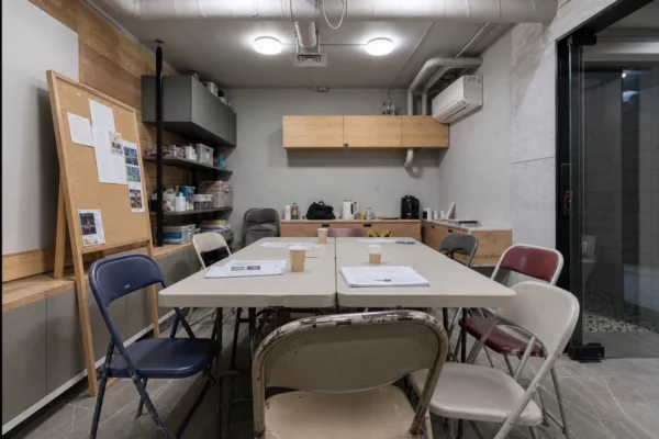 Salle de réunion d’une maison de quartier en France, avec tables pliantes, chaises dépareillées et matériel associatif visible.