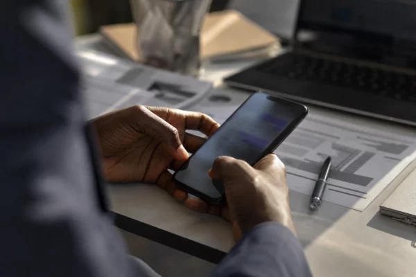 Des mains tiennent un smartphone au-dessus de feuilles imprimées sur un bureau sobre, avec stylo et ordinateur flous en arrière-plan.