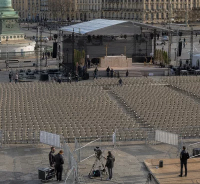 Vue en plongée de rangées de chaises devant une estrade provisoire sur la place de la Concorde, avec façades parisiennes au loin.