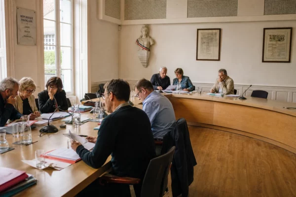 Salle municipale française avec élus anonymes, micros, dossiers flous et une chaise vide en réunion locale