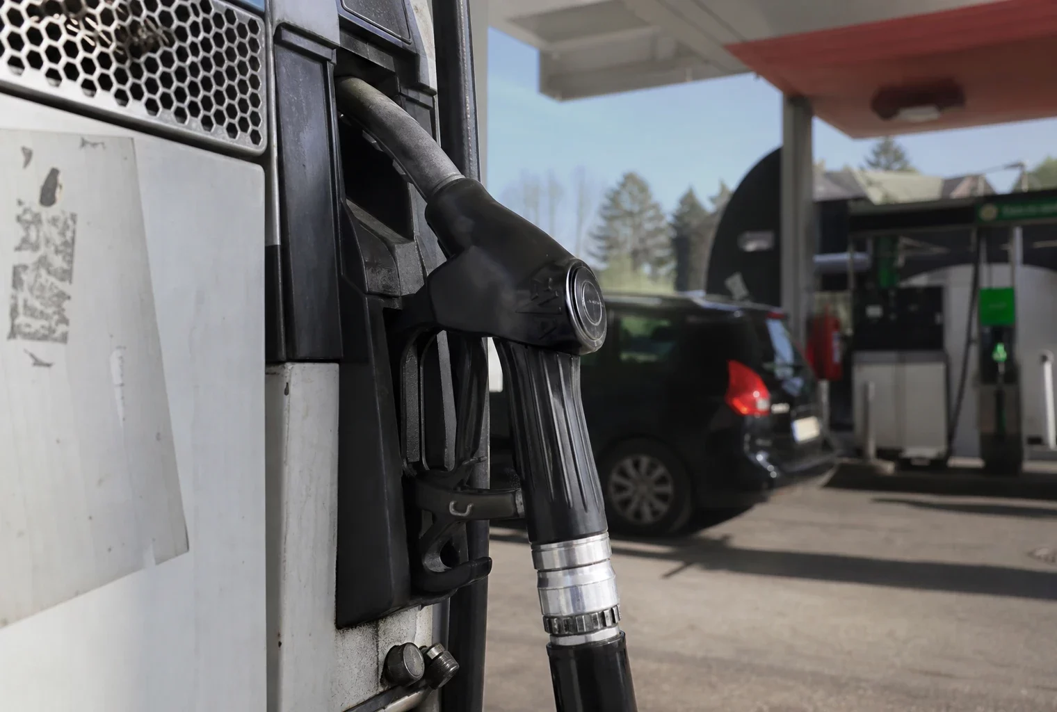 Une pompe à carburant en station-service, avec une voiture en arrière-plan et le prix affiché sur la borne.