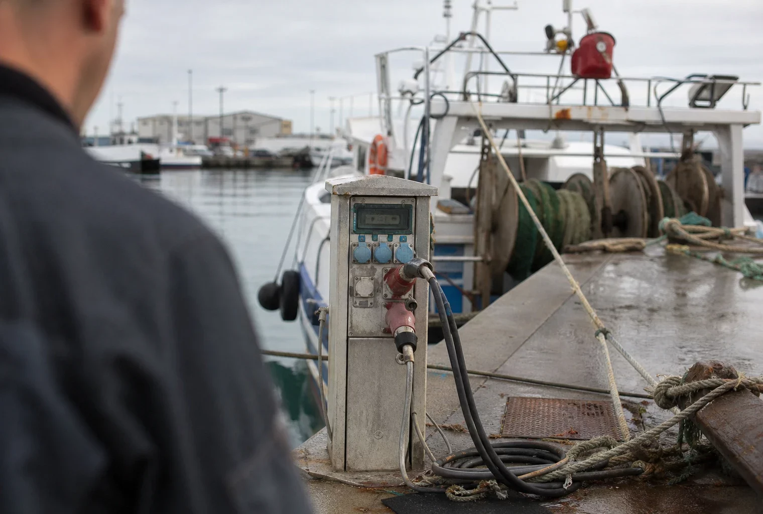 Dans un port de pêche, une borne d’alimentation à quai et des câbles près de bateaux amarrés.