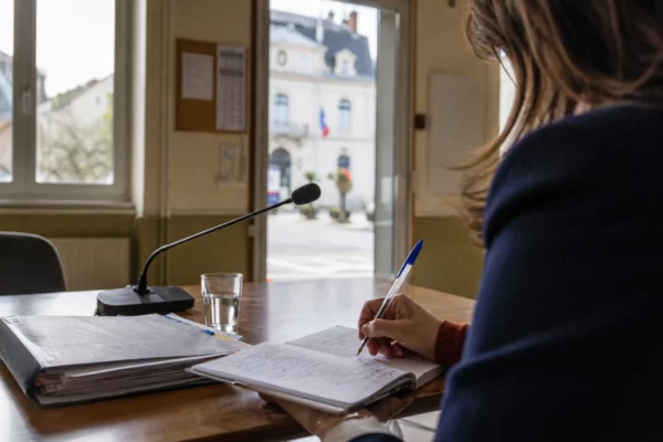 Collaborateur municipal anonyme prenant des notes dans une mairie française, avec un dossier local et une rue en arrière-plan