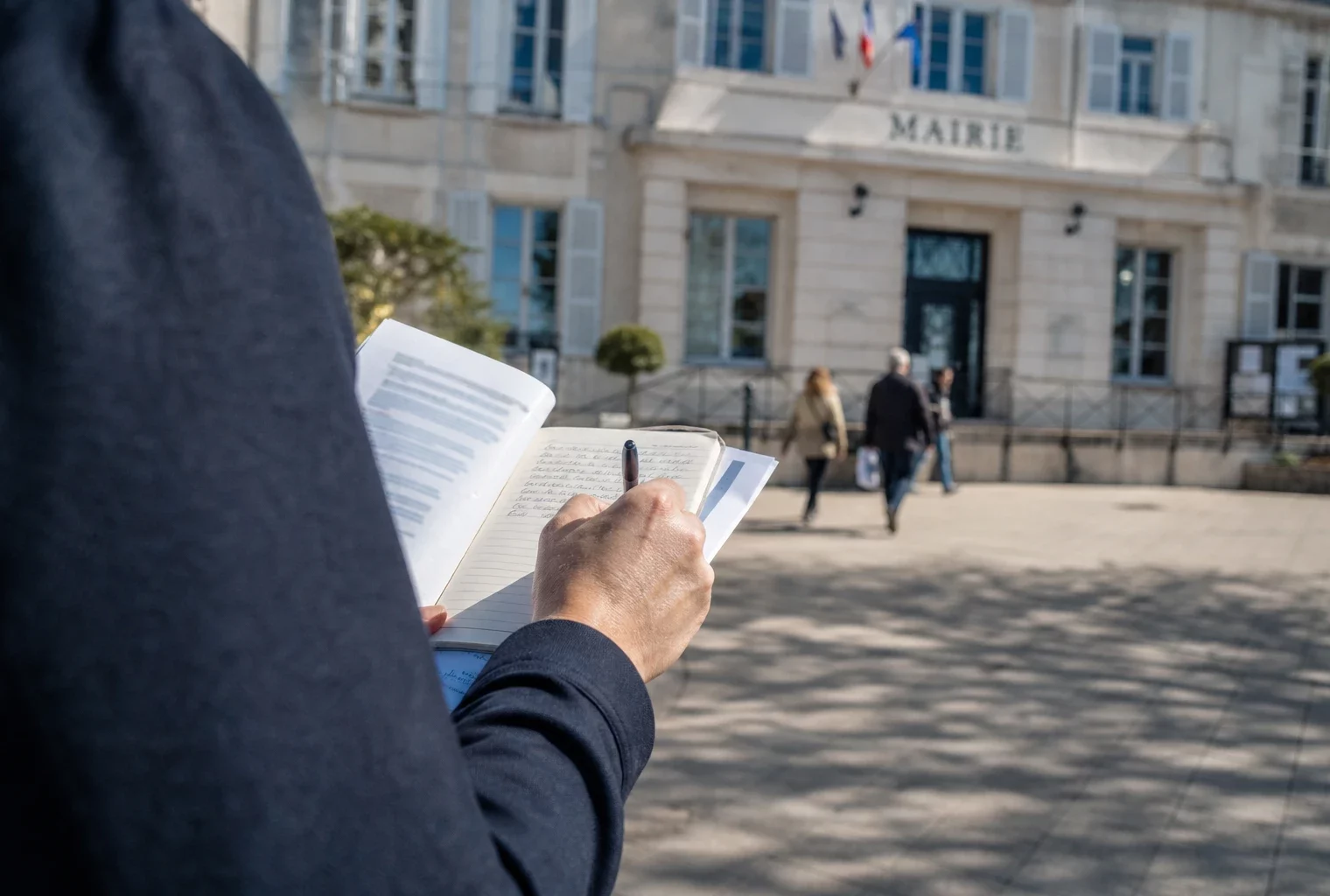 Élu local anonyme prenant des notes devant une mairie française, dans une lumière naturelle de fin de journée