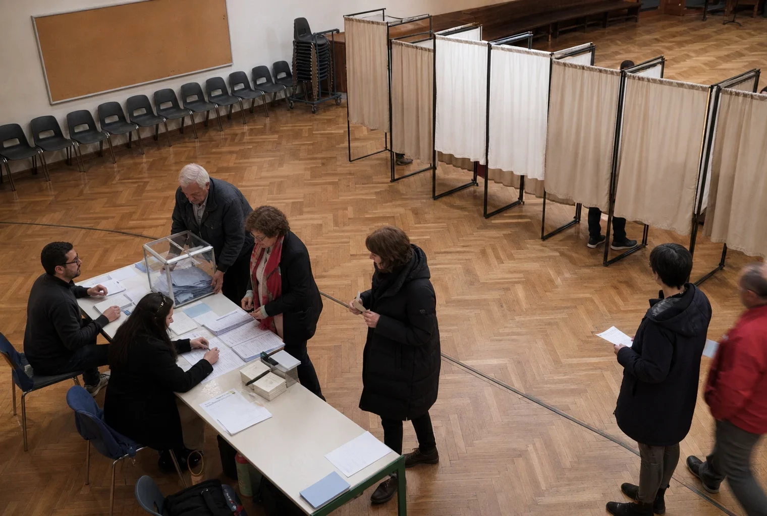 Un bureau de vote de primaire avec isoloirs, table d’émargement et bulletins, dans une salle municipale.