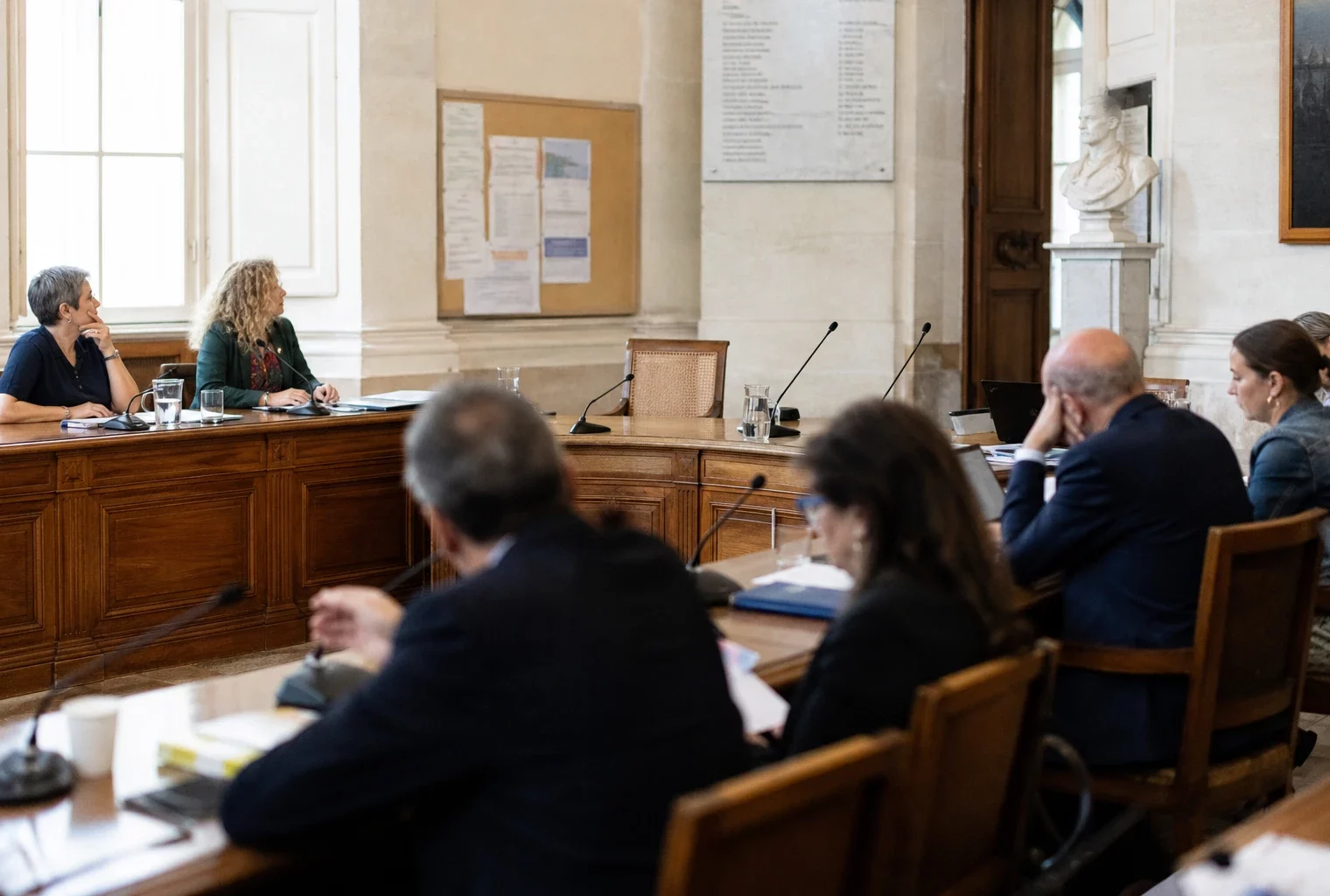 Salle de conseil municipal française avec une chaise vide, des micros et des dossiers avant un débat politique