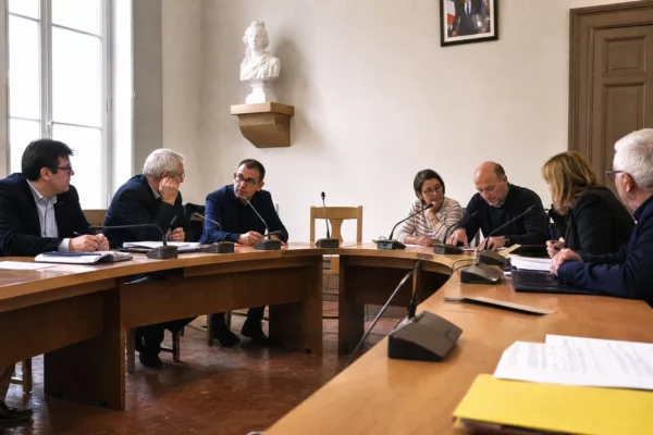 Conseil municipal français en réunion, avec une chaise vide, des micros et des dossiers autour d’une table