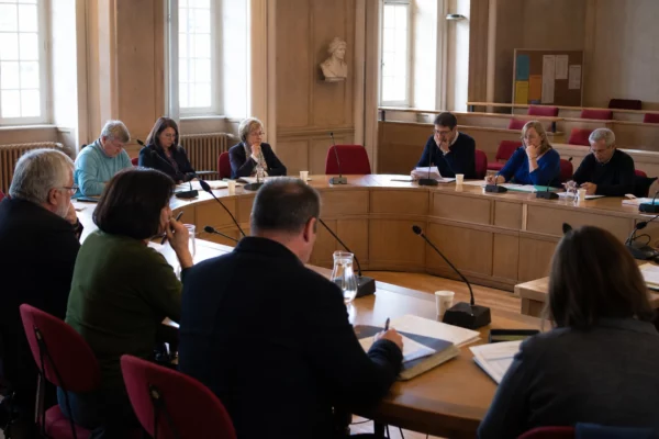 Salle municipale française réunissant des élus autour de micros, dossiers flous et chaise vide en période de primaire politique